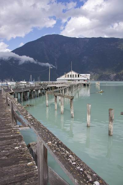 Dilapidated wharf building with rotted wood decking and wood piles in Bella Coola at meeting point of glacial blue Bella Coola River and the sea with the mountains and low cloud behind in British Columbia in Canada Northern America