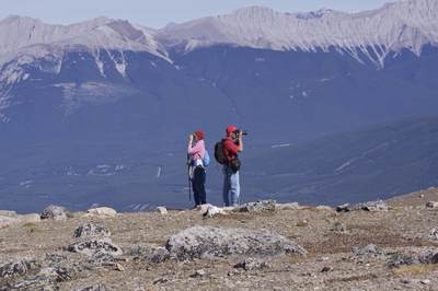 A man and a woman wearing red hats and hiking gear try to capture the Rockies from the summit of Whistlers Mountain on a sunny day with mountains and forest in the distance near Jasper in Alberta in Canada in North America